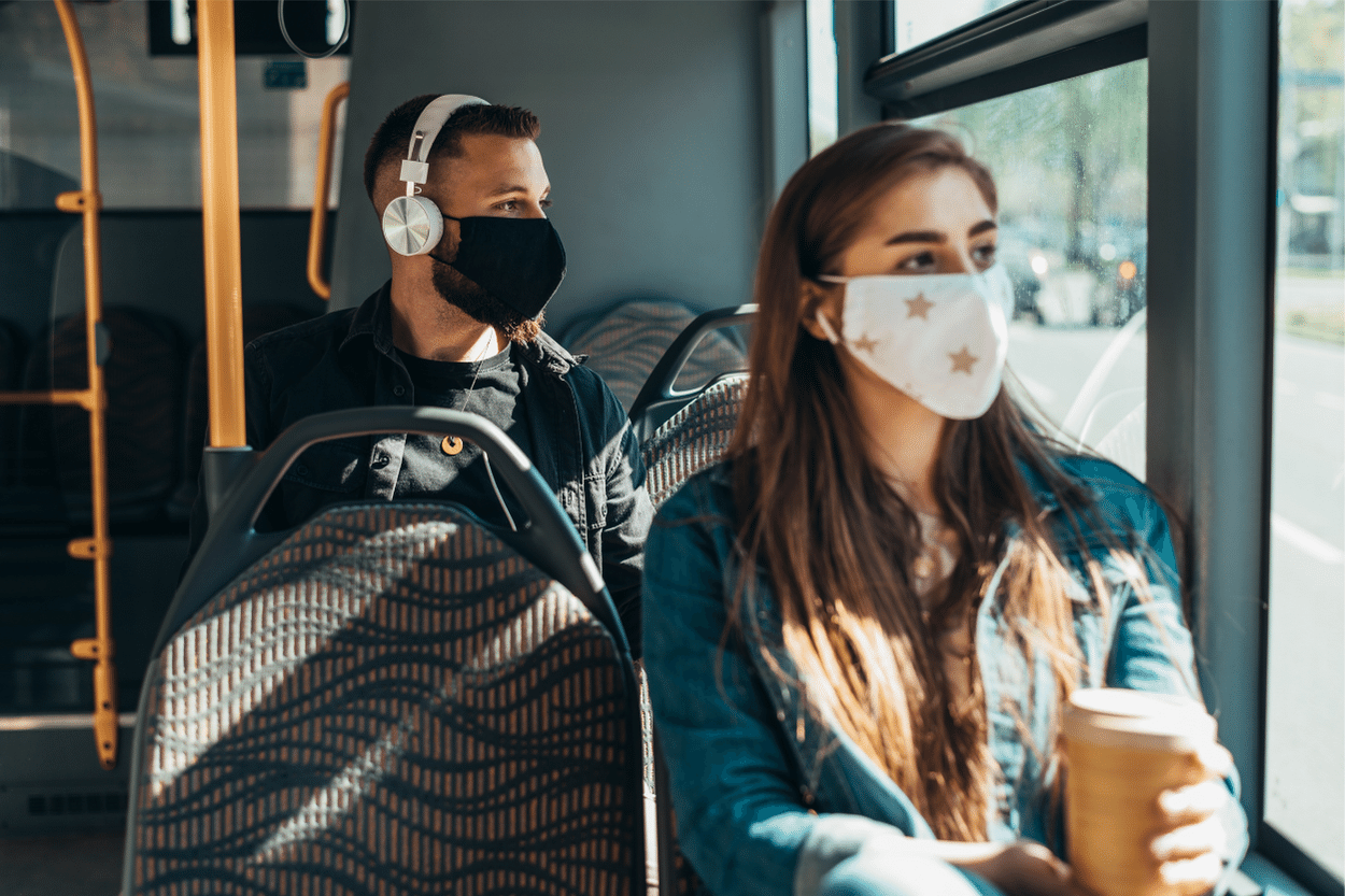 Young woman holding coffee and riding on public transportation and young man in the row behind her in a face mask with headphones on